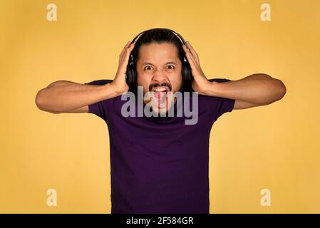 Young man screaming and listening music using headphones and playing with the smartphone. Yellow background. Stock Photo