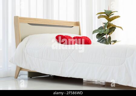 Red heart shaped pillow on bed in bedroom with white curtains, Valentine's day couple concept. Stock Photo
