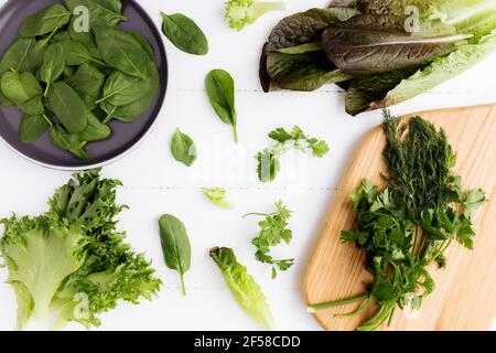 Flat lay with cutting board and bowl of fresh green salad leaves of spinach and lettuce, romaine and parsley, basil on white background. Healthy veget Stock Photo