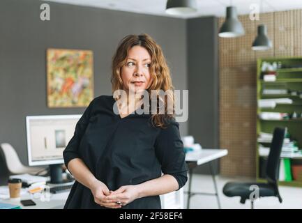 Charming smiling adult woman middle aged woman with curly hair designer manager working in modern office Stock Photo