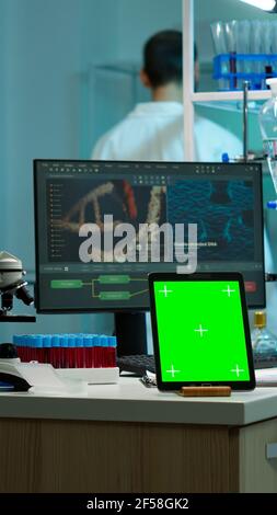 Microbiologist working on monitor in background analysing test samples with tablet with green chroma key screen placed on desk in front. Team of biotechnology scientists developing drugs. Stock Photo