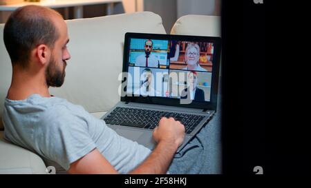 Over shoulder view of man in pajamas having online conversation with teammates working at online technology project using laptop computer, videoconference web internet communication Stock Photo
