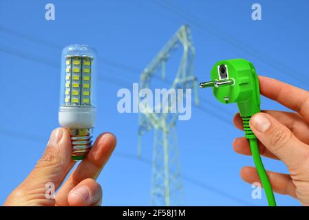Hand holding eco LED light bulb and green electric plug with transmission tower at the background. Energy saving concept. Stock Photo