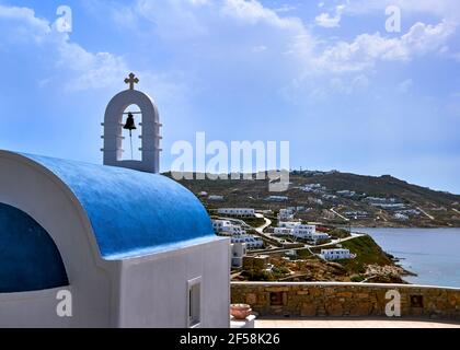 Traditional Greek Orthodox church, blue dome, bell tower overlooking typical Greek island landscape at summer day. Green hills, calm harbor, blue sky. Stock Photo