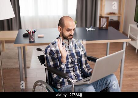 Entrepreneur in wheelchair communicating online with business partners. Businessman waving during video conference, working remotely from home on laptop. Stock Photo