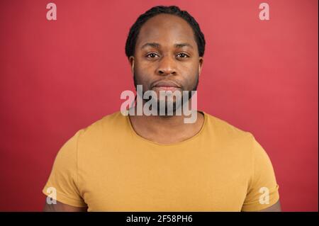 Portrait of calm young handsome African American guy in yellow t-shirt standing isolated over red background and confidently looking at the camera, good looking serious real person black man Stock Photo