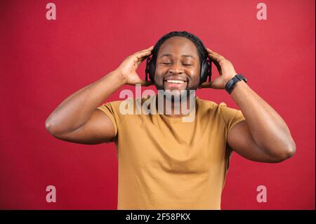 Studio portrait of relaxed cheerful African American man wearing and holding wireless headphones isolated on red wall, enjoying listening to favorite music soundtrack, wellbeing, meditation concept Stock Photo