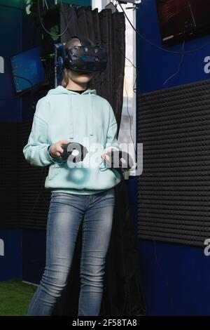 Young teenage girl in virtual reality helmet tries to control game using controllers in VR club Stock Photo