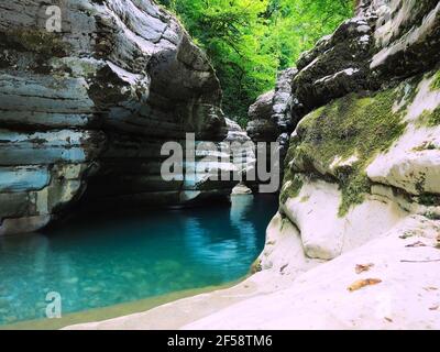 A bright turquoise calm river flows between mossy rocks with bright green trees at the top. Stock Photo