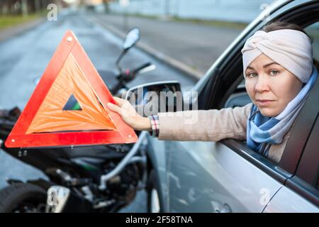 Woman a driver showing triangle emergency sign while sitting in a car after road accident with motorcycle, does not leave the car Stock Photo
