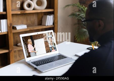 Multiracial male student studying online, watching webinar holding female teacher, other students involved. An African-American man member of video conference, using laptop for connecting online Stock Photo