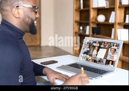 Diverse work team discussing business strategy online, back view over shoulder of African male employee at laptop screen with video profiles of colleagues on it, virtual meeting concept Stock Photo