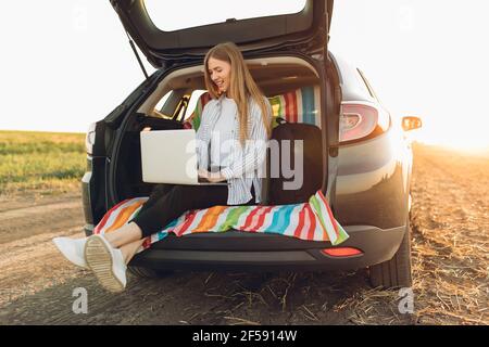 Business attractive young woman working on laptop while sitting in trunk of car while traveling Stock Photo