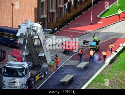 Overhead view of workers laying red tarmac or red asphalt or red bitumen or red tar to create a pavement on a new road in the Brunswick area of Ardwick, Manchester, England, United Kingdom. Workers use rakes to level the tarmac as a small road roller waits to crush it into a smooth surface. A lorry alongside delivers more red asphalt. Stock Photo