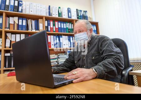 Mature Caucasian man wearing facemask using laptop at work place in office, protection due Covid-19 pandemic Stock Photo