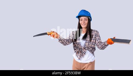 Breaking gender stereotypes. Confident woman in a blue safety helmet holding a board and a hand saw. Studio gray background. Stock Photo