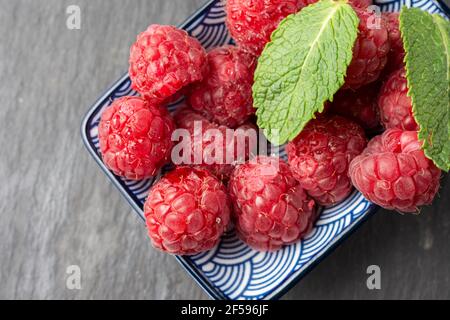 Aerial view of raspberries and mint leaves in bowl, selective focus, on dark wooden table, horizontal Stock Photo