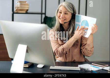 Successful intelligent Asian woman, top manager, ceo, financial tutor, wearing headset, communicates with the business team, via video conference, conducts an online briefing, showing charts, smiling Stock Photo