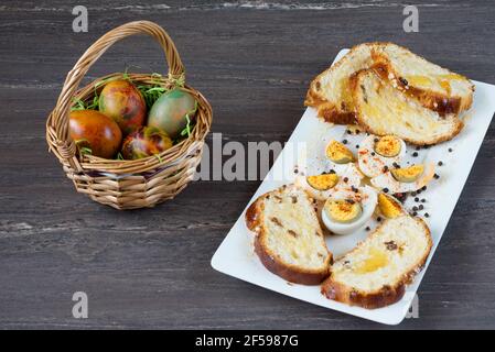 Easter wicker basket with colored eggs and sliced Easter bread in white plate on grey wooden board. Stock Photo