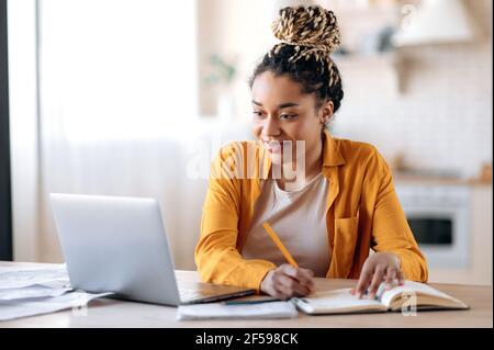Focused cute stylish african american female student with afro dreadlocks, studying remotely from home, using a laptop, taking notes on notepad during online lesson, e-learning concept, smiling Stock Photo