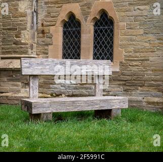 A weathered wooden bench on grass with a stone wall with arched windows in the background. Stock Photo