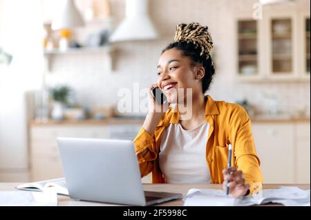African American joyful attractive stylish young woman, freelancer, manager or real estate agent, having pleasant phone conversation with client or employee, sitting at workplace, smiling