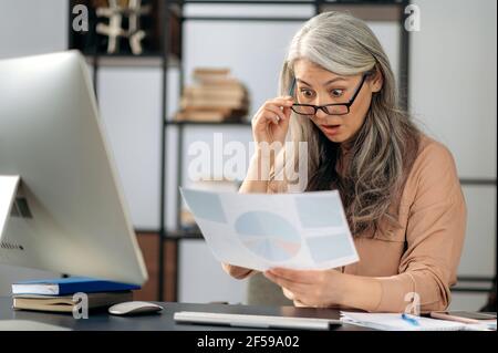Surprised successful senior gray-haired Asian woman, business lady, top manager, sitting at computer, looking at business graphs, studies financial documents with shocked face, taking off glasses Stock Photo