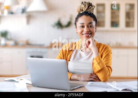 Satisfied good looking young African American stylish woman, freelancer, student or real estate agent, sitting at her desk at home office, looking at the camera and smiling pleasantly Stock Photo