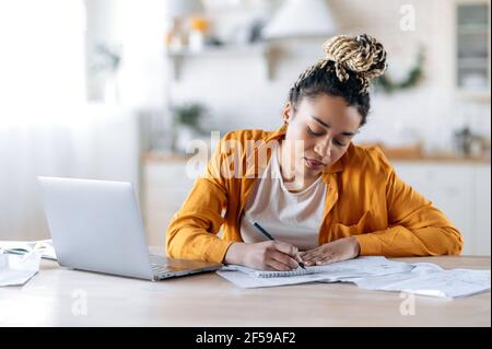 Distance learning, online lesson. Focused smart african american female student, in stylish casual wear, studying remotely, using a laptop, taking notes during online lecture, sitting at home Stock Photo