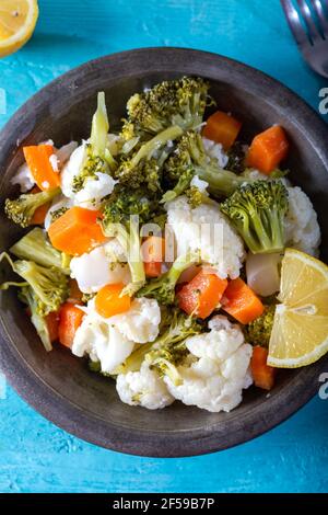 Broccoli, cauliflower and carrot salad in bowl on blue wooden table with olive oil and lemon slices. Top view. Close up. Stock Photo