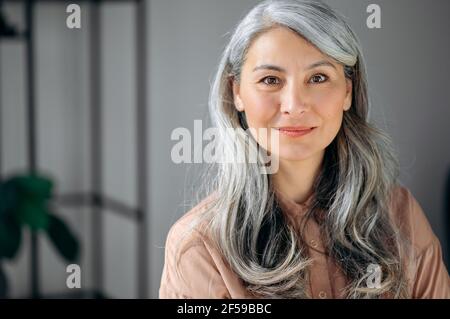 Close-up portrait of beautiful influential confident mature gray-haired Asian woman ceo, lawyer, real estate agent, stands in the office, dressed in stylish clothes, looks at camera, friendly smiling Stock Photo