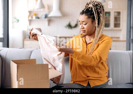 A confused sad african american girl unpacking her parcel at home. Surprised young woman got an unexpected order, girl is upset with her parcel, unsuccessful online shopping. Home delivery Stock Photo