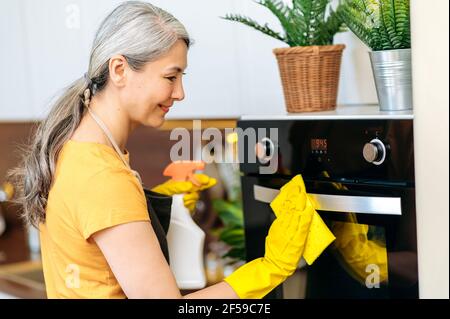 Joyful beautiful hardworking senior gray-haired Asian housewife or cleaning lady in apron and gloves working in kitchen wiping surface of the oven with a rag, holding detergent in her hand, smiling Stock Photo
