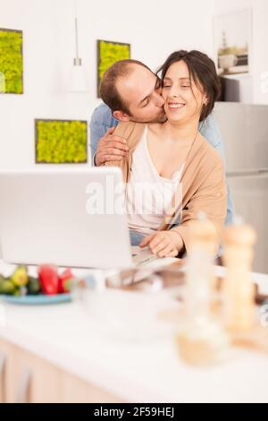Husband making wife smile while kissing her in kitchen. Couple using laptop. Happy loving cheerful romantic in love couple at home using modern wifi wireless internet technology Stock Photo