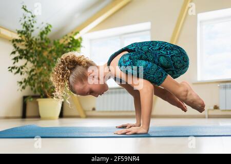 Little beautiful girl with blond hair practicing yoga doing handstand exercise, kakasana or crow pose on a gymnastic mat in the studio. Stock Photo