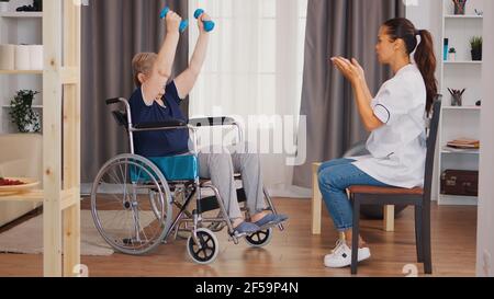 Old lady in wheelchair exercising with dumbbelss. Nurse helping with rehabilitation. Training, sport, recovery and lifting, old person retirement home, healthcare nursing, health support, social assistance, doctor and home service Stock Photo