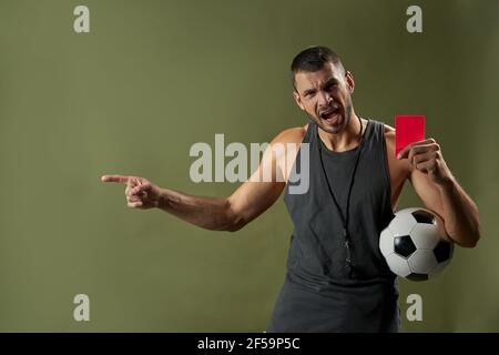 Angry football referee in grey sportswear holding red card in arm isolated on green background of a studio Stock Photo