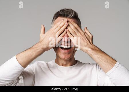 Smiling man covers eyes with hands isolated over gray background Stock Photo