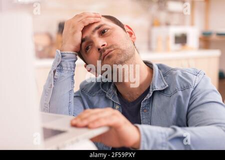 Depressed disabled man in wheelchair using laptop in kitchen. Disabled paralyzed handicapped man with walking disability integrating after an accident. Stock Photo