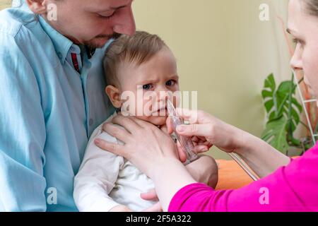 Father holding his baby boy while the mother is using the nasal aspirator to clear the mucus from the nose of her infant. Baby healthcare concept. Stock Photo