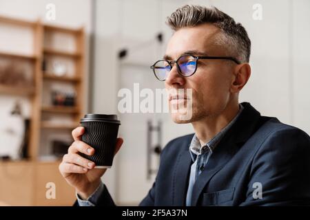 Focused grey man in eyeglasses drinking coffee while working at office Stock Photo