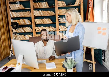 Young adult business partners using laptop, searching ideas for project. Multiracial employees working together in modern office, developing new Stock Photo