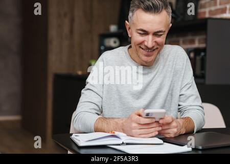 Cheerful handsome man smiling and using mobile phone while working at home Stock Photo