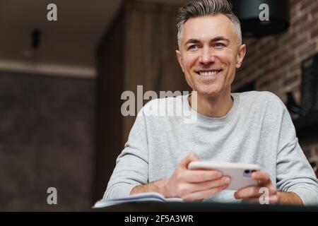 Cheerful handsome man smiling and using mobile phone while working at home Stock Photo