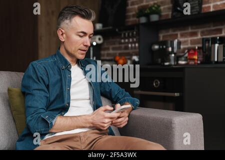 Handsome confident middle aged man using smartphone at home, sitting on a couch Stock Photo