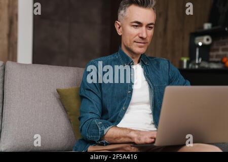 Pleased handsome man using laptop while sitting on couch at home Stock Photo