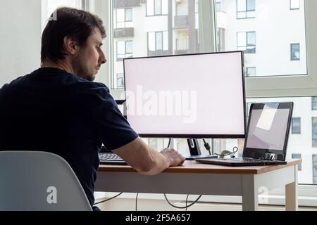 Man is sitting at a workplace with two laptops and a monitor near the window. Remote work from home office Stock Photo