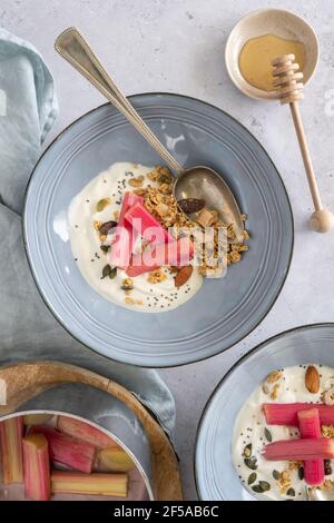 Flat lay of yoghurt and poached rhubarb with granola in two blue bowls with honey, healthy breakfast, vertical on a grey background Stock Photo