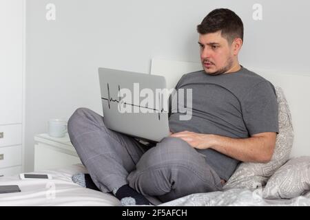 A man works on a laptop sitting in bed, sick, treated, quarantined Stock Photo