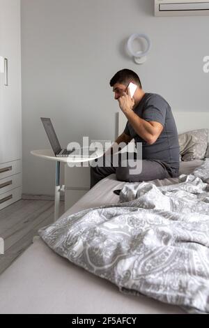 Man talking on the phone, working behind a laptop in the bedroom on th Stock Photo